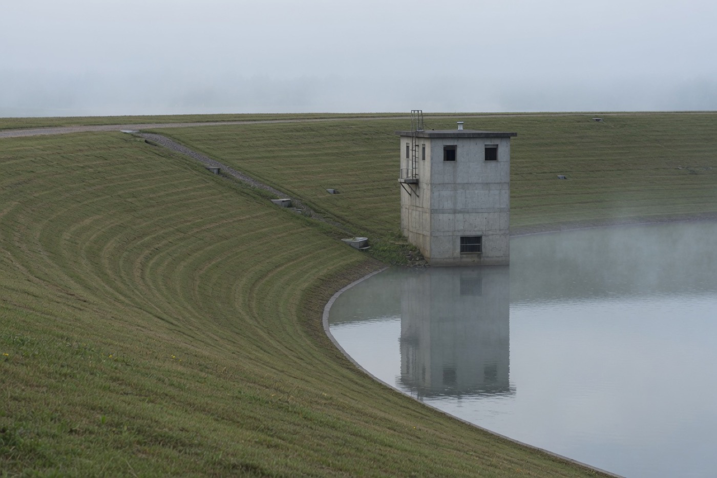 A concrete intake tower at the edge of a reservoir, with a mowed embankment in the mist.
