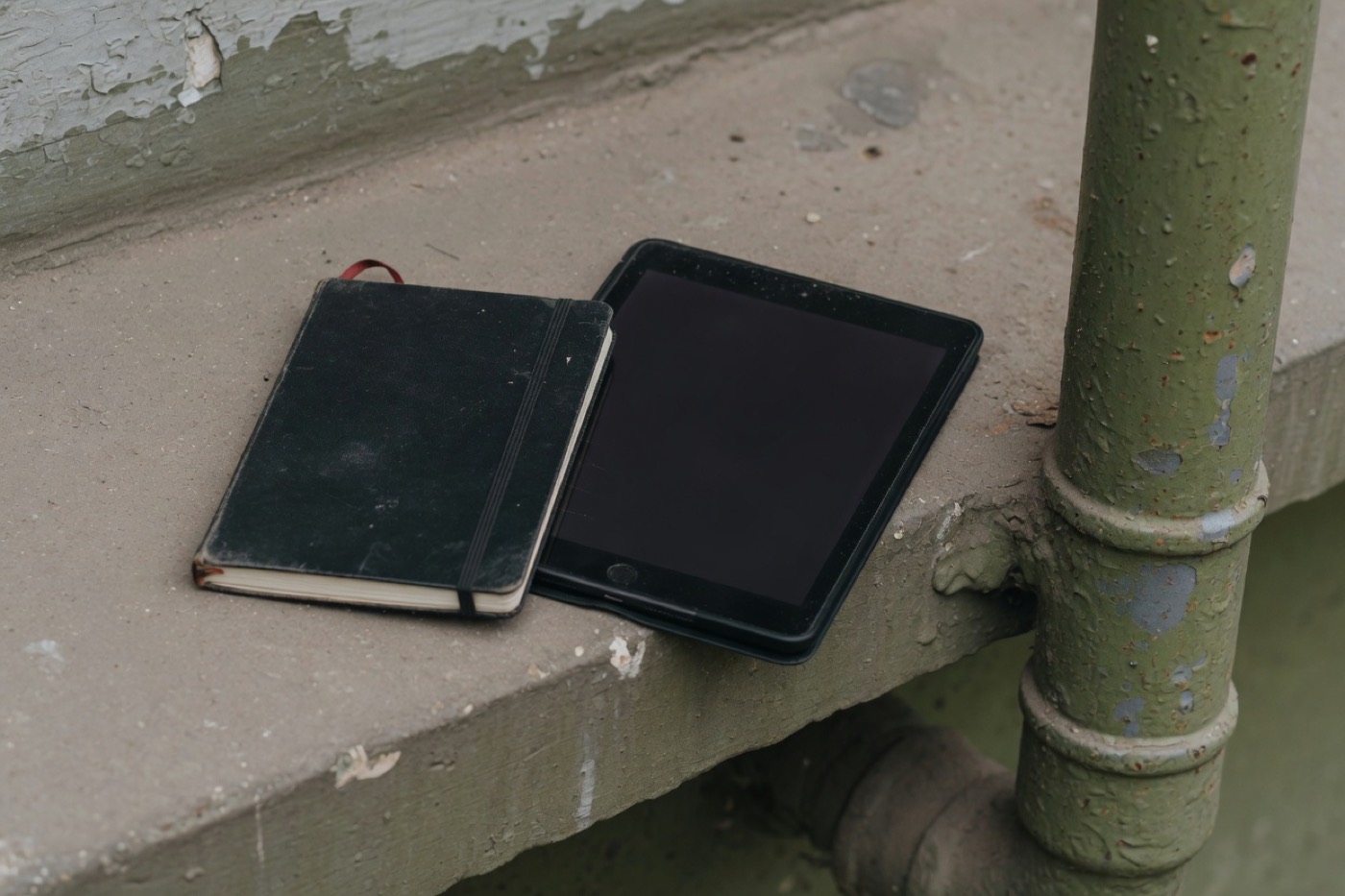 A closed notebook and a tablet on a concrete ledge beside a painted pipe.