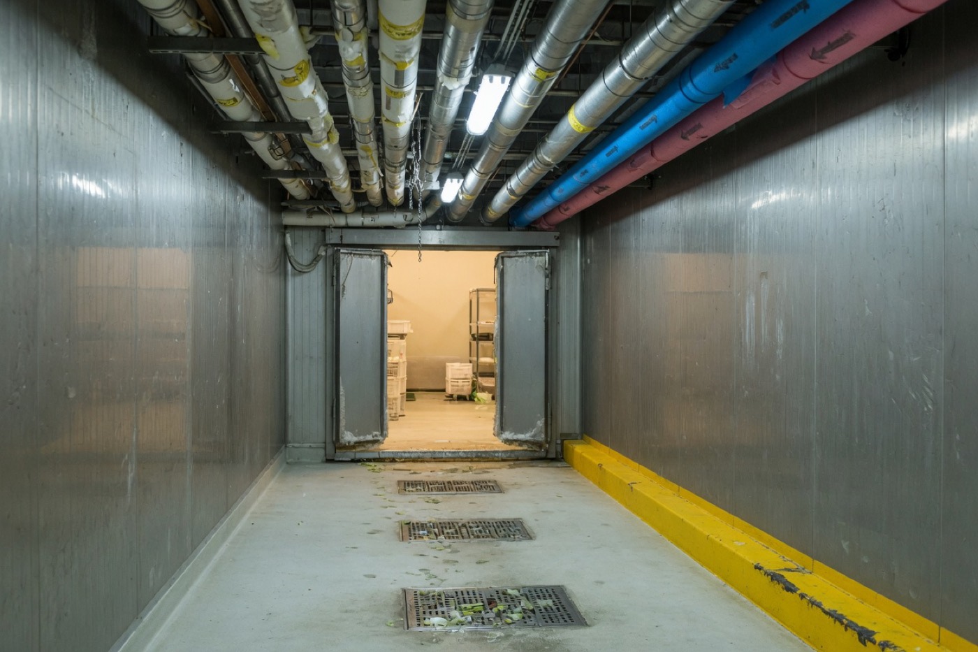 Stainless corridor in a food plant, with floor drains and a warmer room beyond.