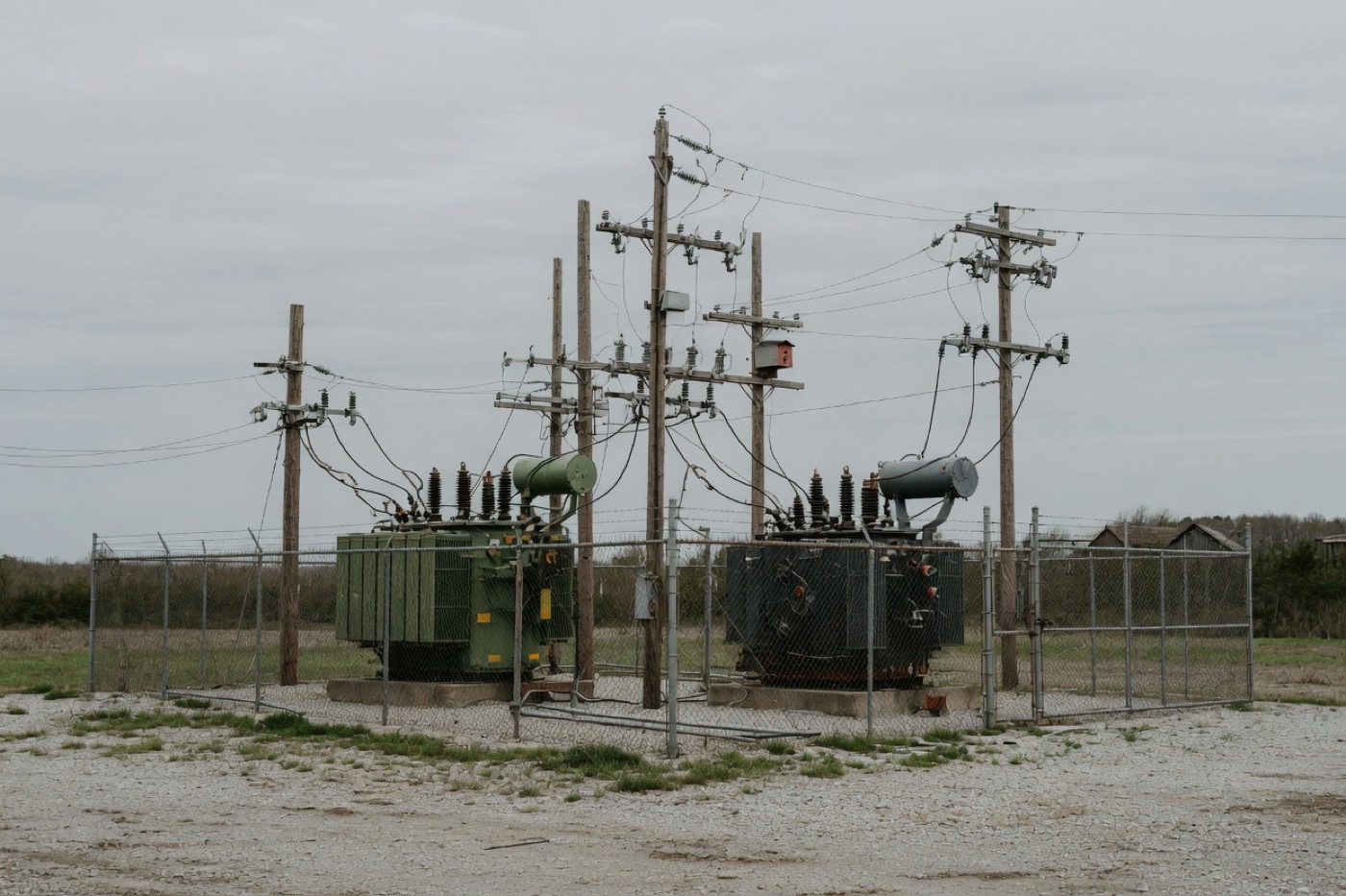 Two transformers in a fenced gravel yard under a gray sky.
